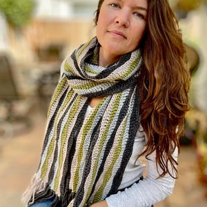 May include: A handmade crochet scarf with vertical stripes in shades of green, gray, black, and beige. The scarf has a fringed edge and is wrapped around the neck. The scarf is a cozy accessory.