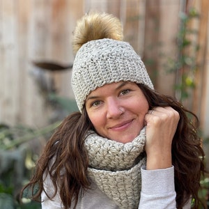 Puede incluir: Una mujer con un gorro de punto gris con un pompón de piel sintética marrón y una bufanda de punto gris a juego.