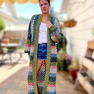May include: A long, open-front crochet cardigan featuring a multi-colored granny square pattern in shades of olive green, dusty pink, cream, and various tones of blue. The garment has long sleeves and a solid olive green ribbed trim along the edges.