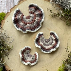 May include: Three crocheted mushroom-shaped decorations in shades of brown, beige, and white. The decorations are arranged on a wooden surface.