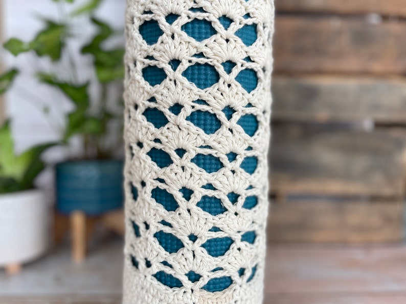 May include: A cream-coloured, crocheted yoga mat carrier. The carrier has an openwork design, revealing a teal-coloured yoga mat inside. The crochet pattern features a repeating fan-like motif. The carrier is cylindrical in shape.