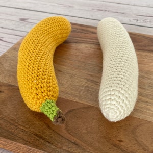 May include: Two crocheted banana toys, one yellow and one white, on a wooden cutting board.