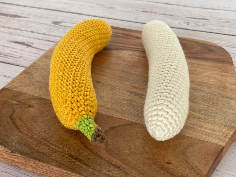 May include: Two crocheted banana toys, one yellow and one white, on a wooden cutting board.