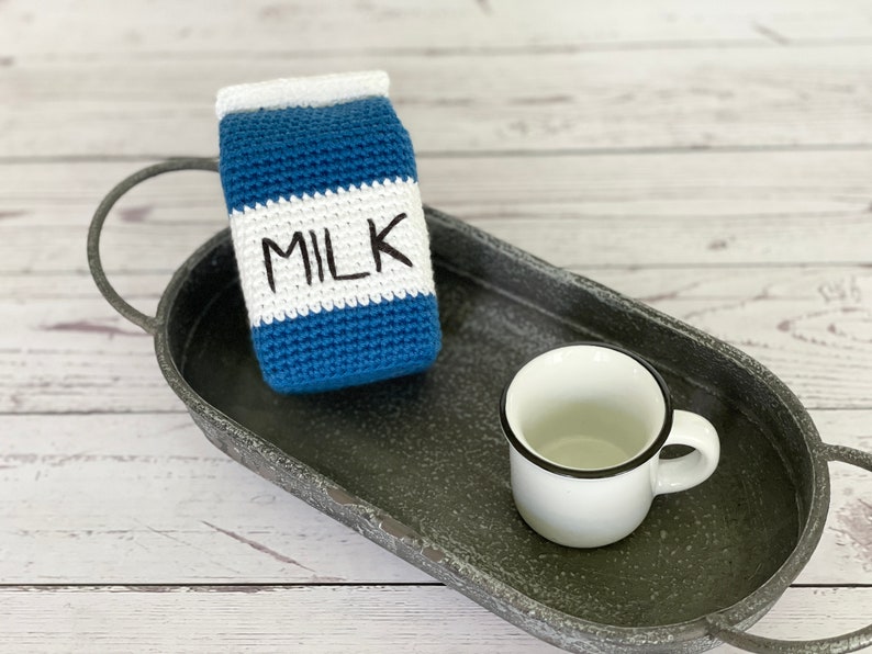 May include: A crocheted milk carton with the word "MILK" on the front, sitting on a gray metal tray with a white ceramic mug.