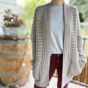 Könnte beinhalten: Ein beigefarbener Häkelcardigan mit langem, offenem Vorderteil und Taschen. Der Cardigan wird über einem grauen T-Shirt und einer burgunderfarbenen Hose getragen.