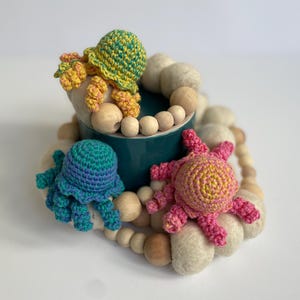 May include: A teal ceramic container surrounded by a felted wool and wooden bead garland. Three crocheted jellyfish in shades of green, blue, and pink are arranged around the container, creating a whimsical, handmade ocean-themed decoration.