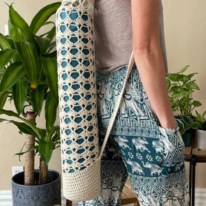 May include: A cream-coloured, crocheted yoga mat bag with a shoulder strap. The bag has an openwork design, revealing a teal yoga mat inside. The bag is cylindrical and appears to be handmade. The person is wearing teal and white patterned trousers.