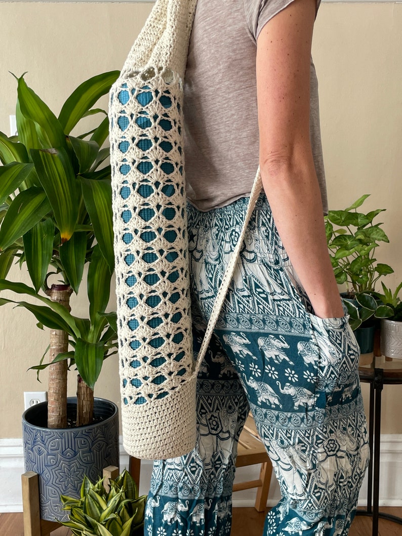 May include: A cream-coloured, crocheted yoga mat bag with a shoulder strap. The bag has an openwork design, revealing a teal yoga mat inside. The bag is cylindrical and appears to be handmade. The person is wearing teal and white patterned trousers.