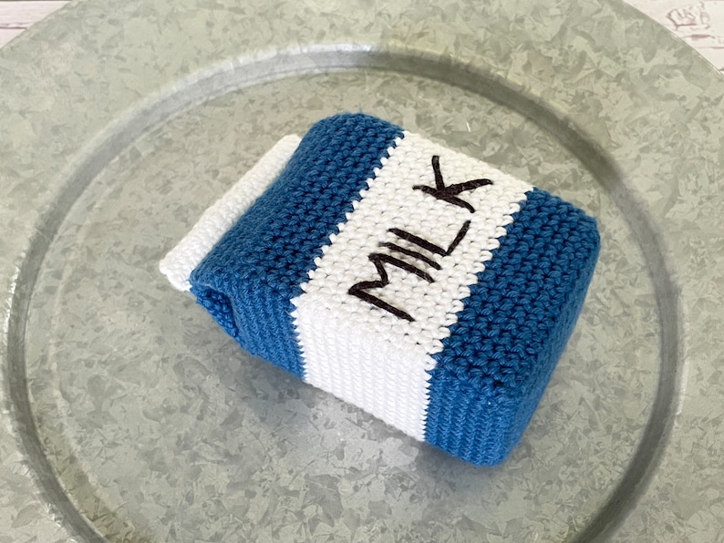 May include: A crocheted milk carton toy with blue and white stripes. The word "MILK" is embroidered in black on the white stripe.