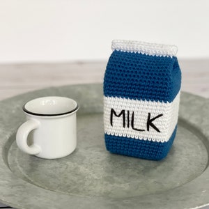 May include: A crocheted milk carton with a white top and a blue body. The carton has the word "MILK" written on it in black. The carton is sitting on a silver tray next to a white mug with a black rim.