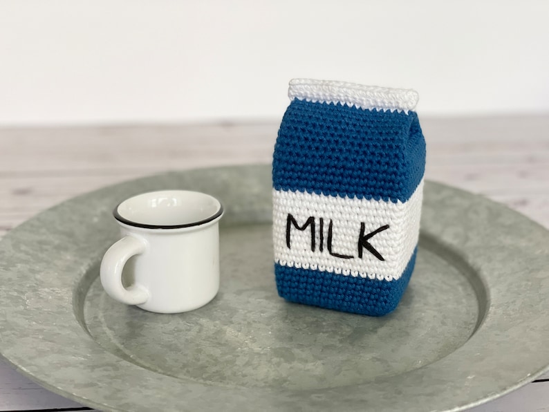 May include: A crocheted milk carton with a white top and a blue body. The carton has the word "MILK" written on it in black. The carton is sitting on a silver tray next to a white mug with a black rim.