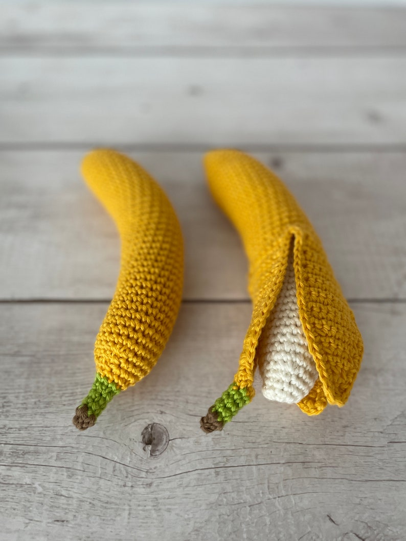 May include: Two crocheted banana toys. One banana is yellow and has a green stem. The other banana is yellow and has a green stem and is partially peeled to reveal a white crocheted inside.