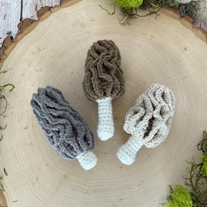 May include: Three crocheted morel mushrooms in different colors: brown, gray, and beige. The mushrooms are sitting on a wooden surface.