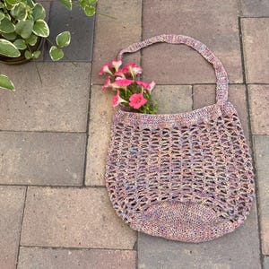 May include: A crocheted mesh bag with a pink, purple, and blue color scheme. The bag has a single strap and is laying on a brick surface with pink flowers inside.