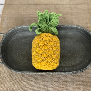 May include: A crocheted pineapple in a grey metal tray. The pineapple is yellow and green with a textured surface.