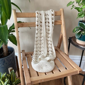May include: A cream-coloured, crocheted bag with an openwork design, draped over a wooden folding chair. The bag features a rounded base and a long strap, highlighting its handmade quality. The chair's natural wood tone complements the bag's neutral colour.