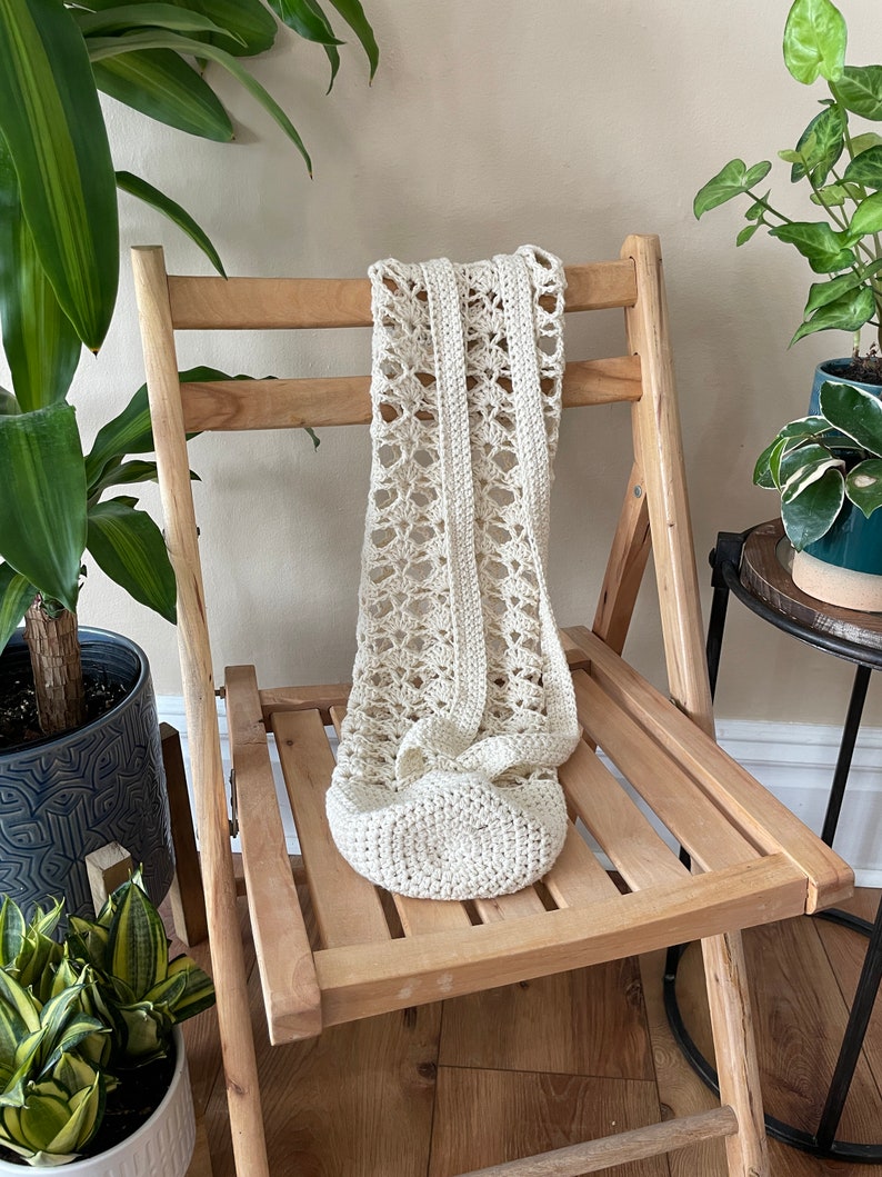 May include: A cream-coloured, crocheted bag with an openwork design, draped over a wooden folding chair. The bag features a rounded base and a long strap, highlighting its handmade quality. The chair's natural wood tone complements the bag's neutral colour.