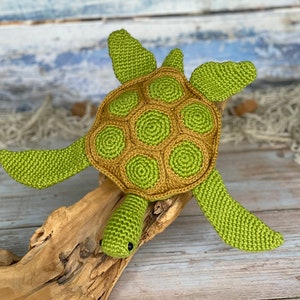 Peut inclure: Une tortue de mer en crochet verte avec une carapace brune. La tortue est assise sur un morceau de bois flotté.