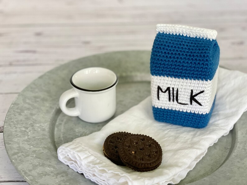 May include: A crocheted milk carton with the word "MILK" on the front, a white ceramic mug, and two chocolate chip cookies on a white napkin.