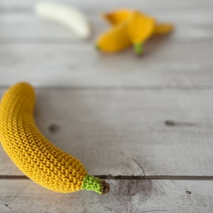 May include: A crocheted yellow banana with a green stem, lying on a white wooden surface.