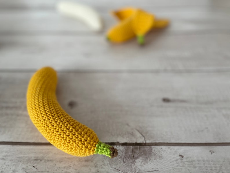 May include: A crocheted yellow banana with a green stem, lying on a white wooden surface.