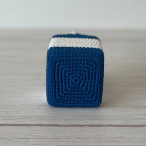 May include: A blue and white crocheted cube toy. The cube is made with a textured stitch pattern and has white stripes around the top.