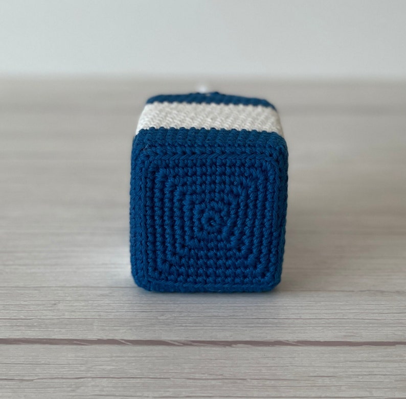 May include: A blue and white crocheted cube toy. The cube is made with a textured stitch pattern and has white stripes around the top.