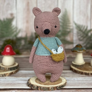 May include: A crocheted pink bear wearing a blue and green shirt and carrying a small basket with two white eggs. The bear is standing on a wooden slice.