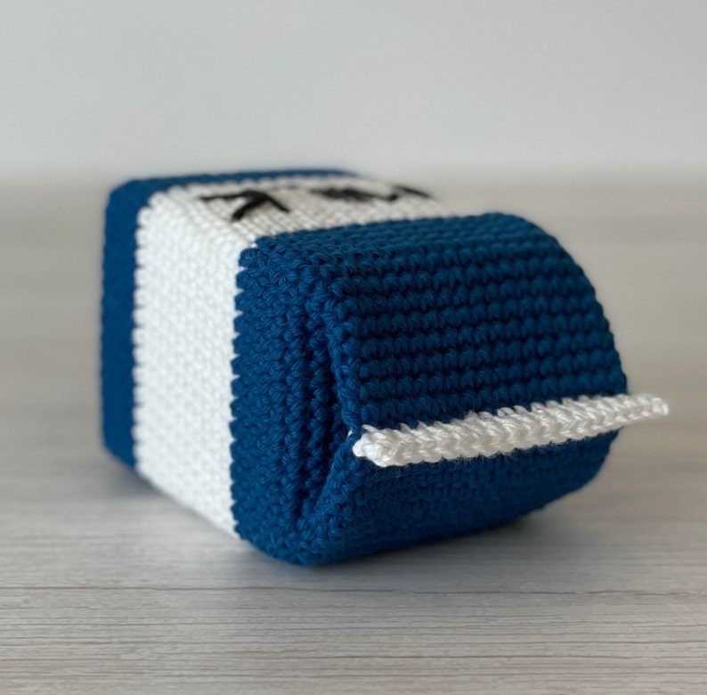 May include: A crocheted milk carton toy with a blue and white striped design. The carton has a white flap that opens to reveal the inside.