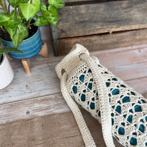 May include: A cream-coloured, open-weave crochet yoga mat bag with a long strap. The bag is cylindrical, with a teal interior visible through the crochet pattern. The bag is lying on a wooden surface with a burlap texture.