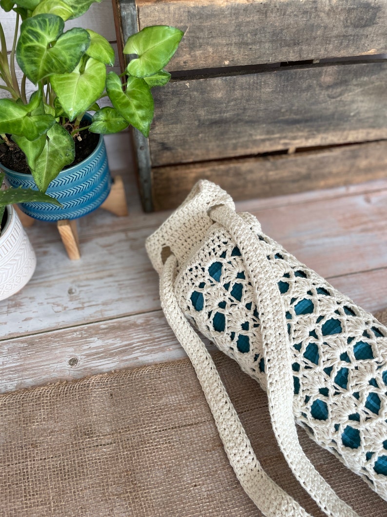May include: A cream-coloured, open-weave crochet yoga mat bag with a long strap. The bag is cylindrical, with a teal interior visible through the crochet pattern. The bag is lying on a wooden surface with a burlap texture.