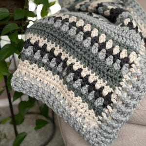 May include: A handmade crochet blanket in shades of grey, cream, and black. The blanket features a classic granny square pattern, with a textured appearance. The soft, cosy blanket is folded over a neutral surface.