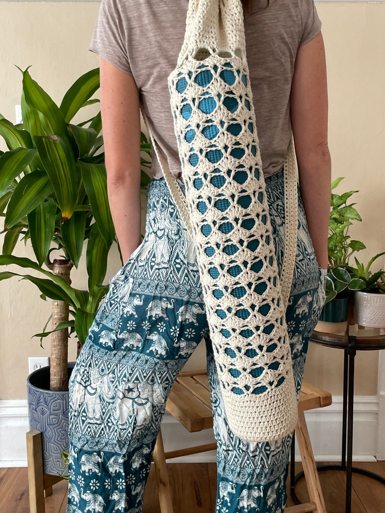 May include: Cream crochet yoga mat bag with a shoulder strap. The bag features an open, geometric design, showing a blue yoga mat. The bag is carried over the shoulder.