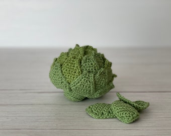 May include: A crocheted artichoke with green leaves and a smaller crocheted artichoke leaf. The artichoke is sitting on a light wood surface.