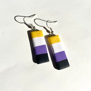 May include: A pair of non-binary pride flag earrings. The earrings are yellow, white, purple, and black. They are made of polymer clay and have silver hooks.