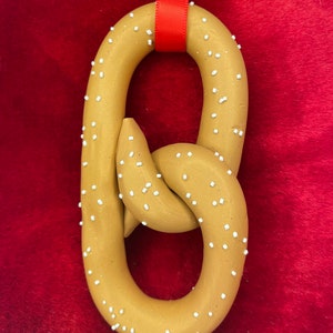 May include: A brown pretzel-shaped Christmas ornament with white sprinkles. The ornament is hanging from a red ribbon.