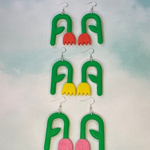 Może przedstawiać: Trzy pary zielonych kolczyków w kształcie kwiatów z różnymi kolorami środka kwiatu. Pierwsza para ma pomarańczowe środki kwiatów, druga para ma żółte środki kwiatów, a trzecia para ma różowe środki kwiatów.