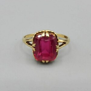 May include: A gold ring with a large, bright red, rectangular gemstone set in a gold bezel. The ring has a delicate, openwork design.