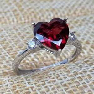 May include: A silver ring with a large heart-shaped red gemstone and two smaller clear gemstones on the band.