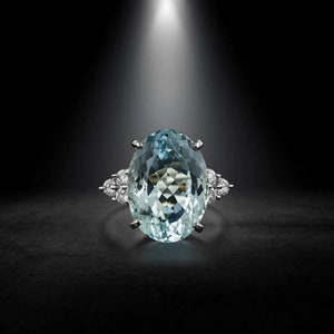 May include: An aquamarine and diamond ring. The ring features a large, oval-cut aquamarine gemstone, surrounded by smaller diamonds. The setting is silver-toned, and the ring is displayed against a dark background.