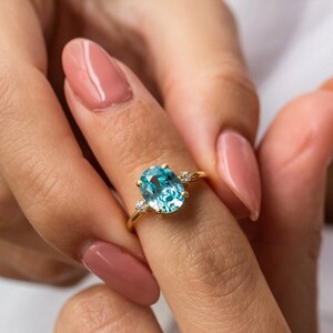 May include: A gold ring with a large oval blue gemstone and two smaller round clear gemstones. The ring is on a finger.