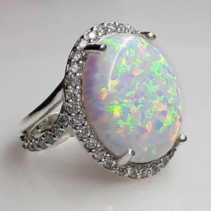 Könnte beinhalten: Silberner Ring mit einem großen, ovalen Opal-Edelstein. Der Opal zeigt ein Farbspiel mit grünen, gelben und violetten Blitzen. Der Edelstein ist von einem Kranz aus kleinen, klaren Kristallen umgeben, was seine Brillanz verstärkt.