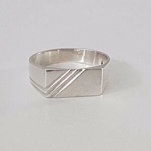 May include: A silver signet ring with a rectangular top and three engraved lines.