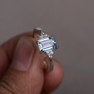 May include: A silver ring with a light blue emerald-cut aquamarine gemstone and smaller clear gemstones on the band.