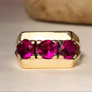 May include: A gold ring with three round, deep pink gemstones set in a rectangular band.