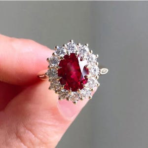 May include: A gold ring with a large, oval-cut, deep red gemstone at its centre, encircled by a halo of smaller, clear gemstones. The ring has a simple gold band. The setting is a classic, elegant design.