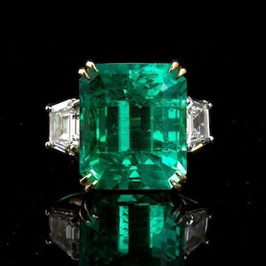 May include: A gold ring featuring a large emerald green gemstone flanked by two smaller, clear gemstones.