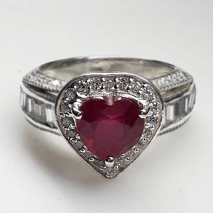 May include: A silver ring with a heart-shaped ruby gemstone surrounded by smaller white gemstones. The ring has a detailed band with a textured design.