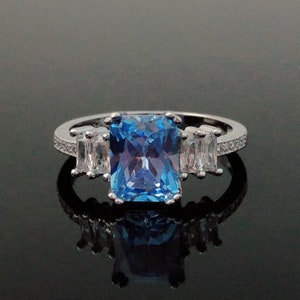 May include: A silver ring with a large, rectangular blue gemstone surrounded by smaller, clear gemstones.