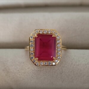 May include: A gold ring with a large, square, red gemstone surrounded by smaller, clear gemstones.